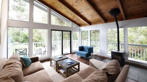 Ein geräumiges Wohnzimmer mit hoher Decke und großen Fenstern, einem gemütlichen Sofa, einem blauen Sessel und einem Kaminofen.