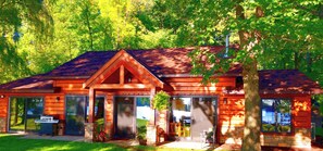 A wooden cabin with a large sliding glass door, a patio with a grill, and a view of the forest.