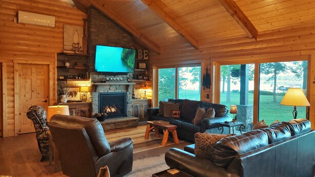 Interior acogedor de una cabaña de troncos con chimenea de piedra, muebles de cuero y un ventanal con vistas al exterior.
