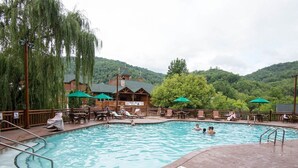A swimming pool with lounge chairs and umbrellas, surrounded by a wooden deck and a building with a green roof. - 2BR with Great Mountain views Master Suite and JR suite (Gatlinburg)