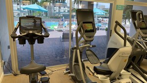 Ein Fitnessraum mit verschiedenen Trainingsgeräten, darunter ein Crosstrainer und ein Fahrradergometer, mit Blick auf einen Pool und Palmen.