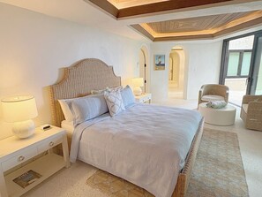 A bedroom with a large bed, a nightstand with a lamp, a chair, and a view of the outdoors.