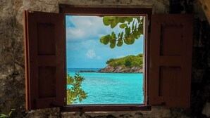 Een raam met houten luiken, met uitzicht op turquoise water en een rotsachtig eiland begroeid met groen.