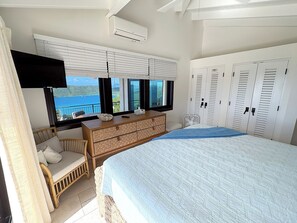 A bedroom with a bed, a dresser, a television, and a view of the sea.