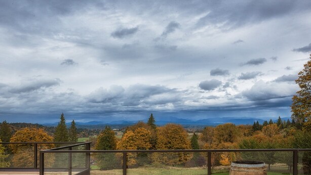 Ein Balkon mit Glasgeländer, der auf eine Landschaft mit Herbstbäumen und entfernten Bergen unter bewölktem Himmel blickt.