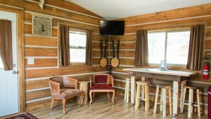 A cabin interior with wooden walls, a dining table, chairs, a television, and a window with curtains.