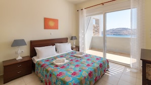 A bedroom with a large bed, two bedside tables with lamps, a view of the sea through a balcony, and a painting on the wall.