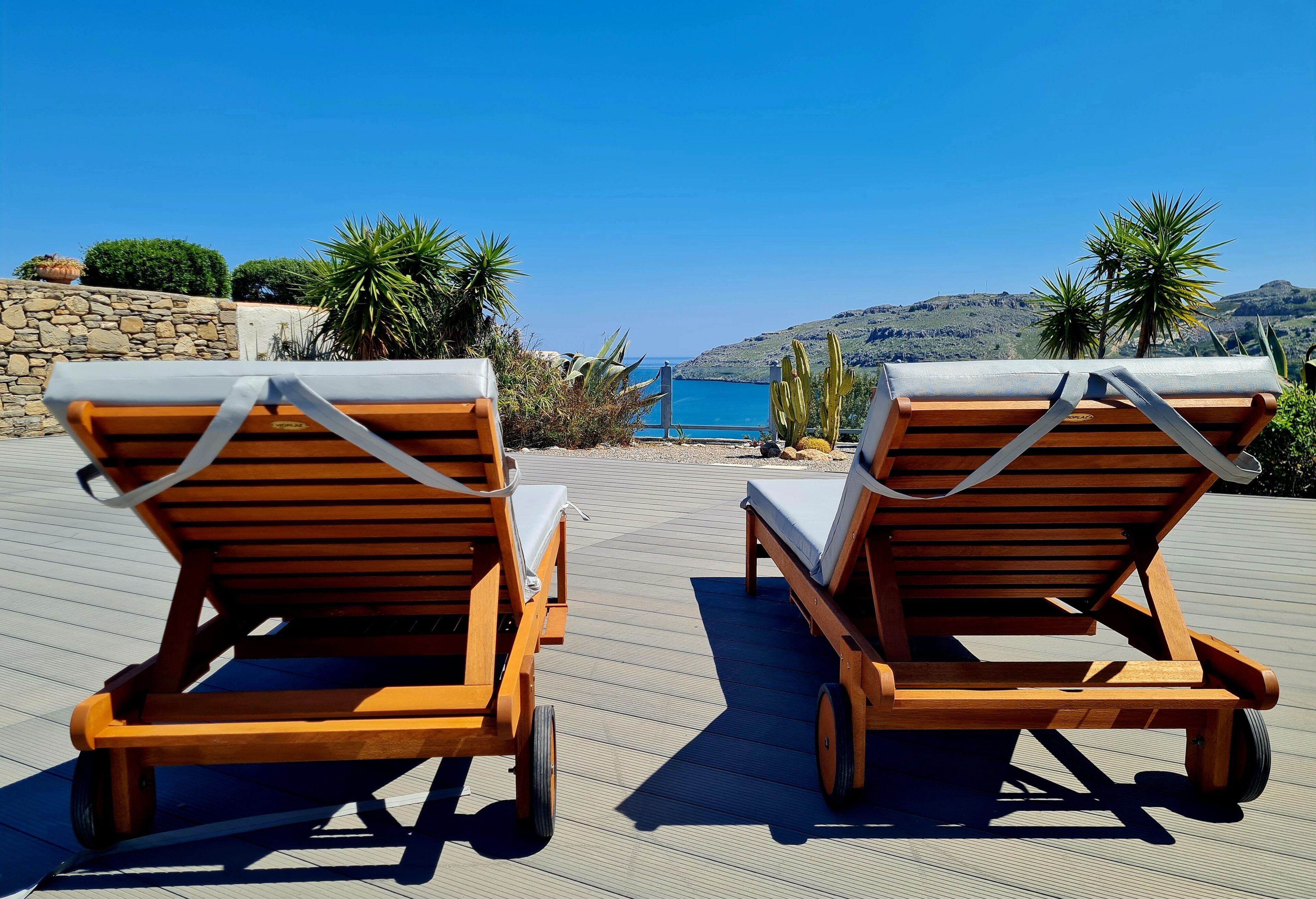 Two wooden lounge chairs with white cushions on a wooden deck overlooking a body of water and hills.