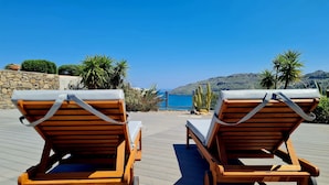Two wooden lounge chairs with white cushions on a wooden deck overlooking a body of water and hills.
