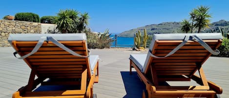 Two wooden lounge chairs with white cushions on a wooden deck overlooking a body of water and hills.