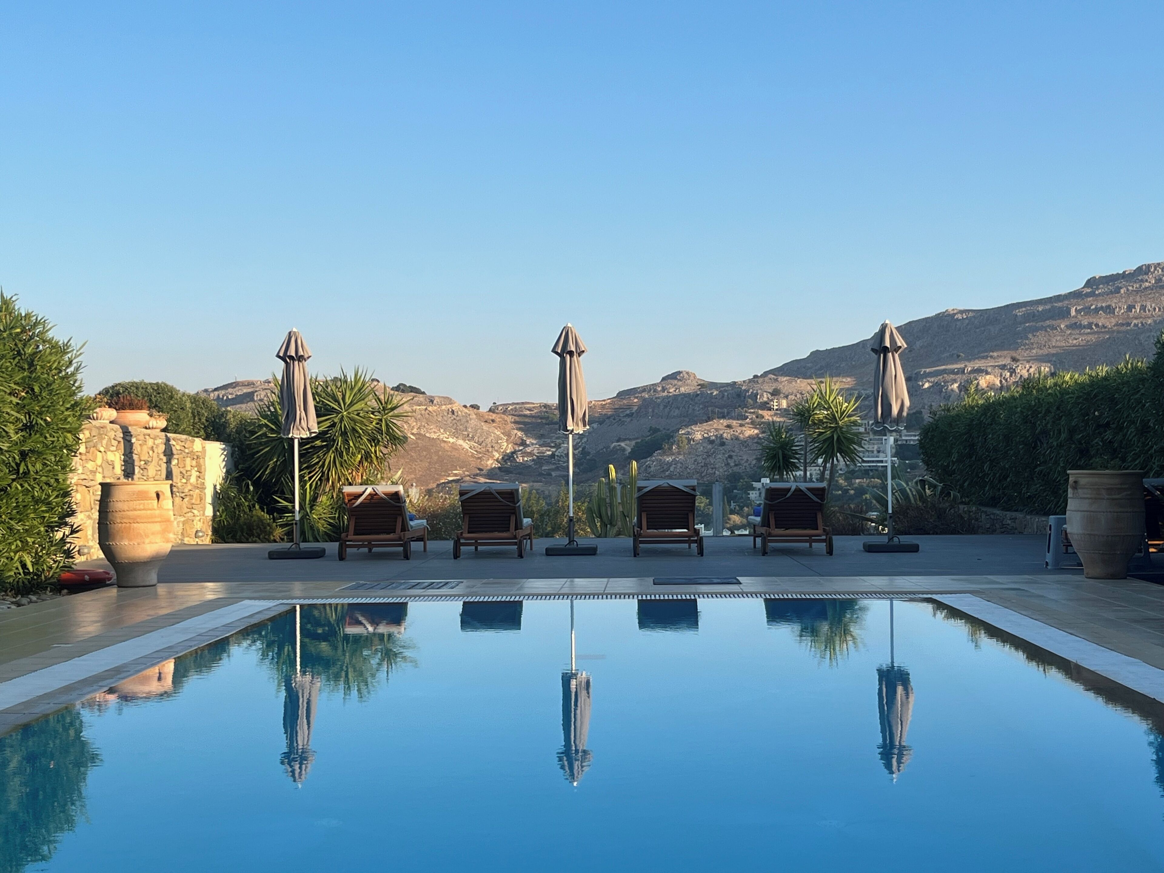A pool with lounge chairs and umbrellas, surrounded by greenery and a mountainous backdrop.