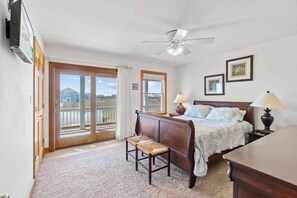 A bedroom with a bed, two bedside tables, a desk, a chair, a ceiling fan, and a view through the glass doors. - Semi-Oceanfront Rodanthe Home w/ Pool, Hot Tub, Elevator, Game and Media Rooms (Rodanthe)