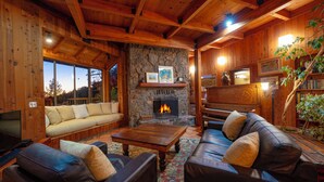 Acogedor interior de cabaña de madera con chimenea de piedra, sofás de cuero y un amplio ventanal con vistas a un paisaje pintoresco.