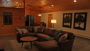 Una sala de estar acogedora con un sofá seccional de cuero marrón, una mesita de centro redonda y una chimenea. Hay cuadros enmarcados en la pared y una lámpara sobre una mesita auxiliar.