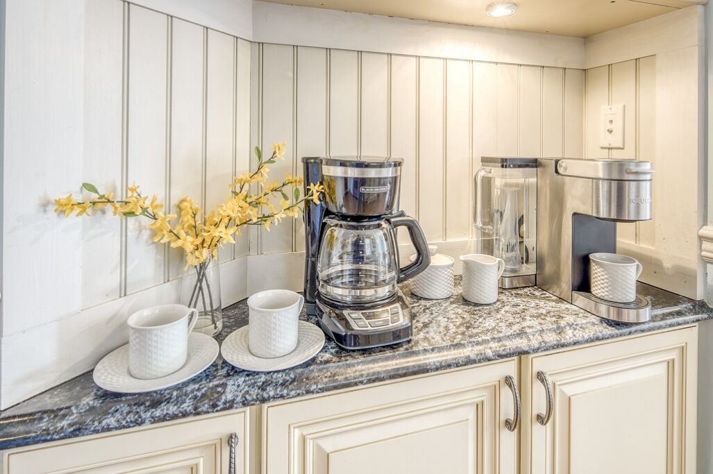 Una cocina con cafetera, hervidor y un florero con flores amarillas sobre la encimera.