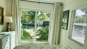 A bedroom with a bed, a nightstand, a lamp, a window with blinds, and a view of a pool and palm trees. - Deep Water Canal, Swim Spa Pool, Kayaks, Walk to Ocean Front Bars & Restaurants! (Marathon)