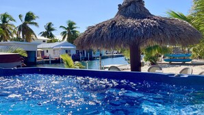 A clear blue pool with a thatched umbrella, surrounded by palm trees and a dock with boats. - Deep Water Canal, Swim Spa Pool, Kayaks, Walk to Ocean Front Bars & Restaurants! (Marathon)