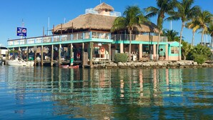 A waterfront building with a thatched roof, a covered deck, and a small boat docked nearby. - Deep Water Canal, Swim Spa Pool, Kayaks, Walk to Ocean Front Bars & Restaurants! (Marathon)