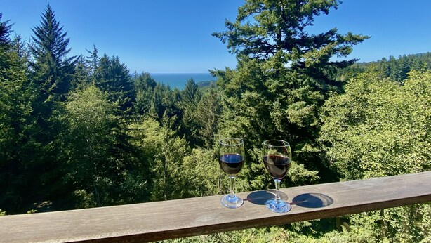 A wooden deck with two wine glasses and a bottle, overlooking a forest and the ocean. - Stunning Custom Cabin in the woods; Mountain and Ocean Views! (Brookings)