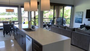 A modern kitchen with a central island, a dining area with chairs, a sofa, and a large window offering a view of the outdoors. - Location Location Location!Ekahi 32C!Designer Remodel 2bd Remaining Fall $400/nt (Wailea-Makena)