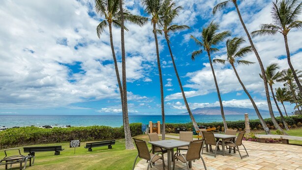 A patio area with a table and chairs, surrounded by palm trees and a view of the ocean. - Location Location Location!Ekahi 32C!Designer Remodel 2bd Remaining Fall $400/nt (Wailea-Makena)