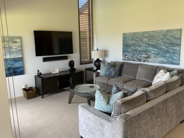 A living room with a grey sofa, a coffee table, a TV, and artwork on the walls. - Location Location Location!Ekahi 32C!Designer Remodel 2bd 2 bth. Ocean View! (Wailea-Makena)
