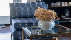A modern living room with a blue sofa, a glass coffee table with a vase of dried flowers, and a bookshelf in the background. - Stunning views in premier winery area. Very private with deck! (Paso Robles)
