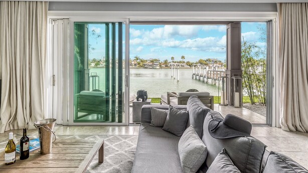 Ein modernes Wohnzimmer mit einer großen grauen Couch, einer Glastür, die auf einen Balkon führt, und Blick auf einen Yachthafen mit Booten und Palmen.