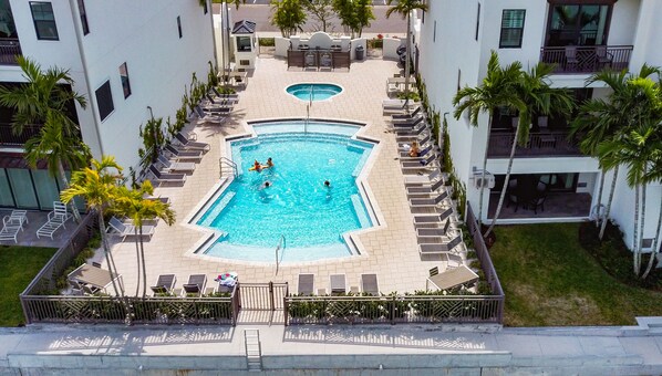 Vista aérea de una piscina rodeada de sillones de descanso y palmeras, con una piscina más pequeña en el centro y una escalera que baja al área de la piscina.