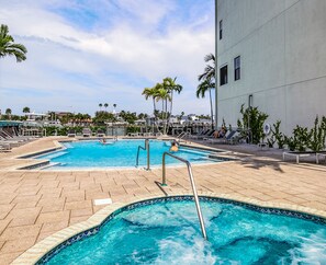 Ein Pool mit gefliestem Rand, umgeben von Liegestühlen und Palmen, mit einem Gebäude im Hintergrund.