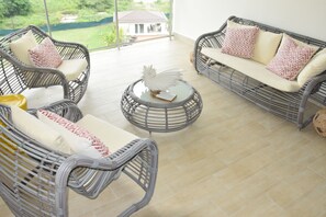 Relax in a spacious room with a high ceiling, featuring a comfortable seating area and a large window offering a view of a lush garden.