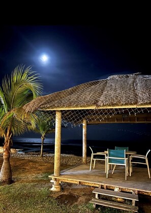 Relax in a serene coastal setting beneath a thatched-roof pavilion with a bench and chairs.