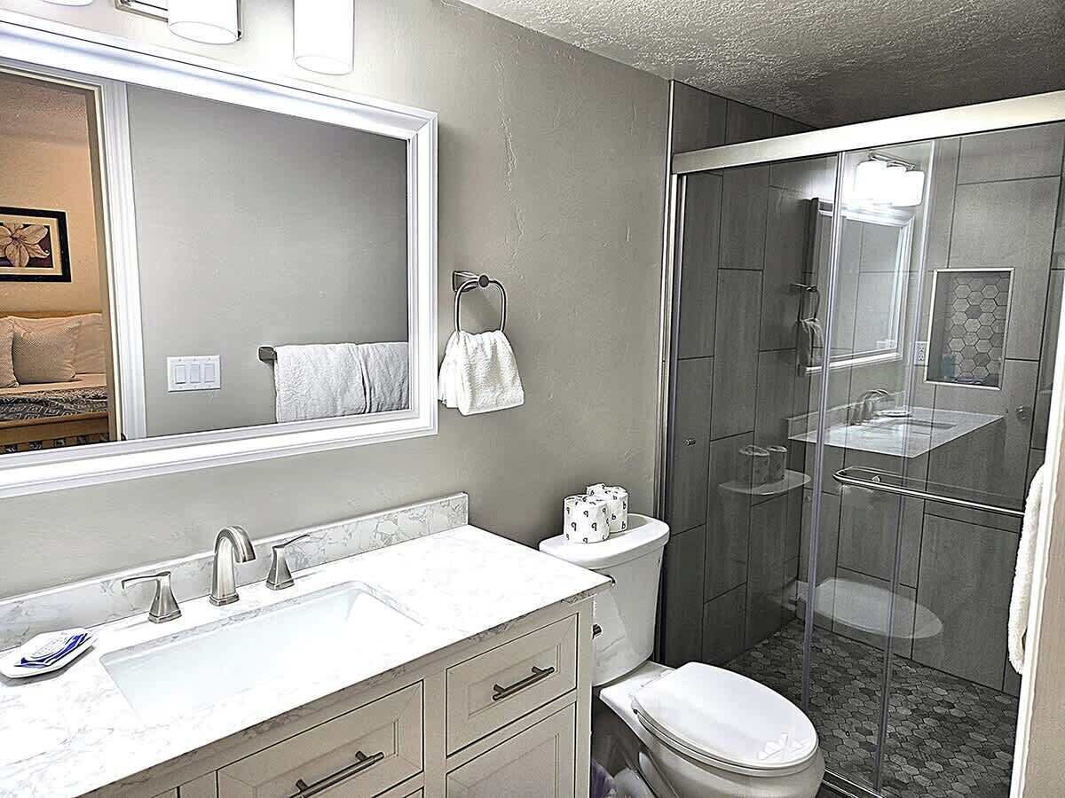 A modern bathroom with a large mirror, a sink with a marble countertop, a toilet, and a glass-enclosed shower area.