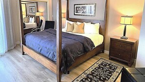 A four-poster bed with a dark blue bedspread, a wooden nightstand with a lamp, and a framed picture on the wall.