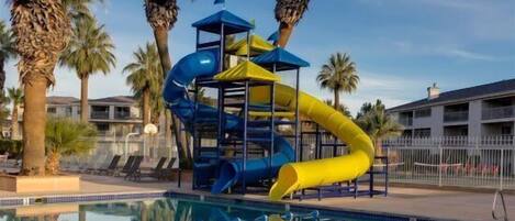 Zona de piscina con tobogán, palmeras y una pileta.
