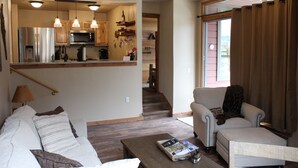 Una sala de estar con sofá, una mesa de centro y una silla. Hay una zona de cocina con refrigerador, microondas y utensilios colgando. Una escalera conduce a otra habitación.