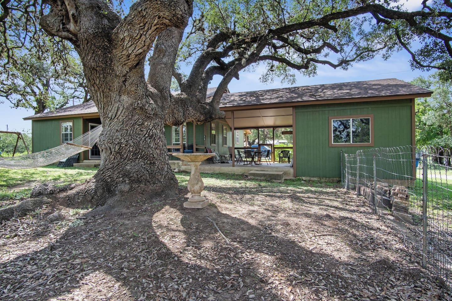 Ein Haus mit einem großen Baum davor, einer Terrasse mit Gartenmöbeln und einem umzäunten Bereich.
