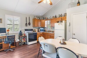 Une cuisine compacte avec des meubles en bois, une cuisinière, un four à micro-ondes et un réfrigérateur. On y trouve une table à manger avec des chaises, un petit espace bureau avec un bureau et un ordinateur, ainsi qu’une fenêtre avec des stores.