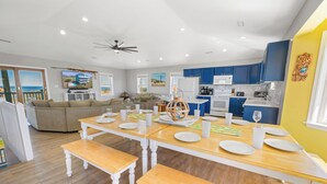 A modern kitchen with blue cabinets, a dining area with wooden tables and benches, and a living room with a sofa and a TV displaying a beach scene. - Oceanview Dog Friendly w/ Private Pool, Hot Tub, Wet Bar. Shore Beats Workin' (Frisco)
