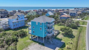 A coastal residential area with houses, including a prominent blue two-story building with a balcony, surrounded by greenery and a clear view of the ocean. - Oceanview Dog Friendly w/ Private Pool, Hot Tub, Wet Bar. Shore Beats Workin' (Frisco)