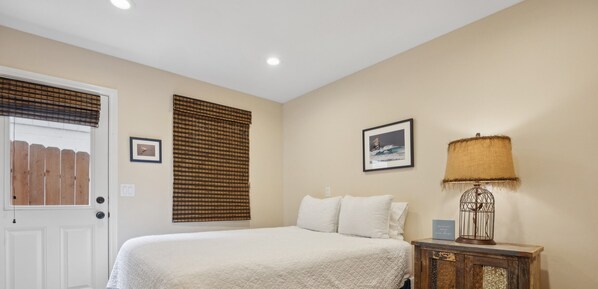 A bedroom with a bed, a wooden door, a window with blinds, a wall-mounted lamp, and two framed pictures. - Beautiful Custom home, great ocean views! Pets Allowed! (Cayucos)