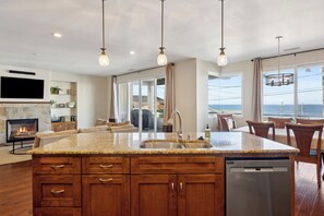 A modern kitchen with granite countertops, wooden cabinets, and a built-in dishwasher. There is a large island with a sink, and the room has large windows offering a view of the ocean. - Beautiful Custom home, great ocean views! Pets Allowed! (Cayucos)