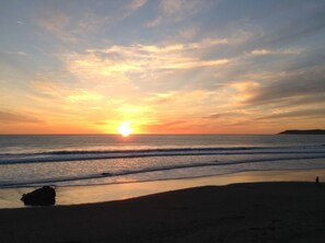 Une plage au coucher du soleil, avec le soleil qui disparaît à l’horizon, les vagues qui viennent doucement lécher le rivage, et la silhouette d’une personne qui se promène.