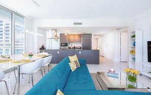 Eine moderne Küche mit Kochinsel, ein Essbereich mit weißem Tisch und Stühlen sowie ein Wohnbereich mit blauem Sofa.