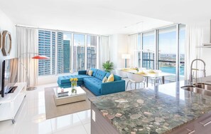 A modern living room with a blue sofa, a dining area, and a kitchen with a large window.