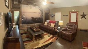 A living room with a leather couch, a coffee table, a TV, and a large wall mural. - Cozy Cabin in the Woods - Lake Livingston, Texas (Livingston)