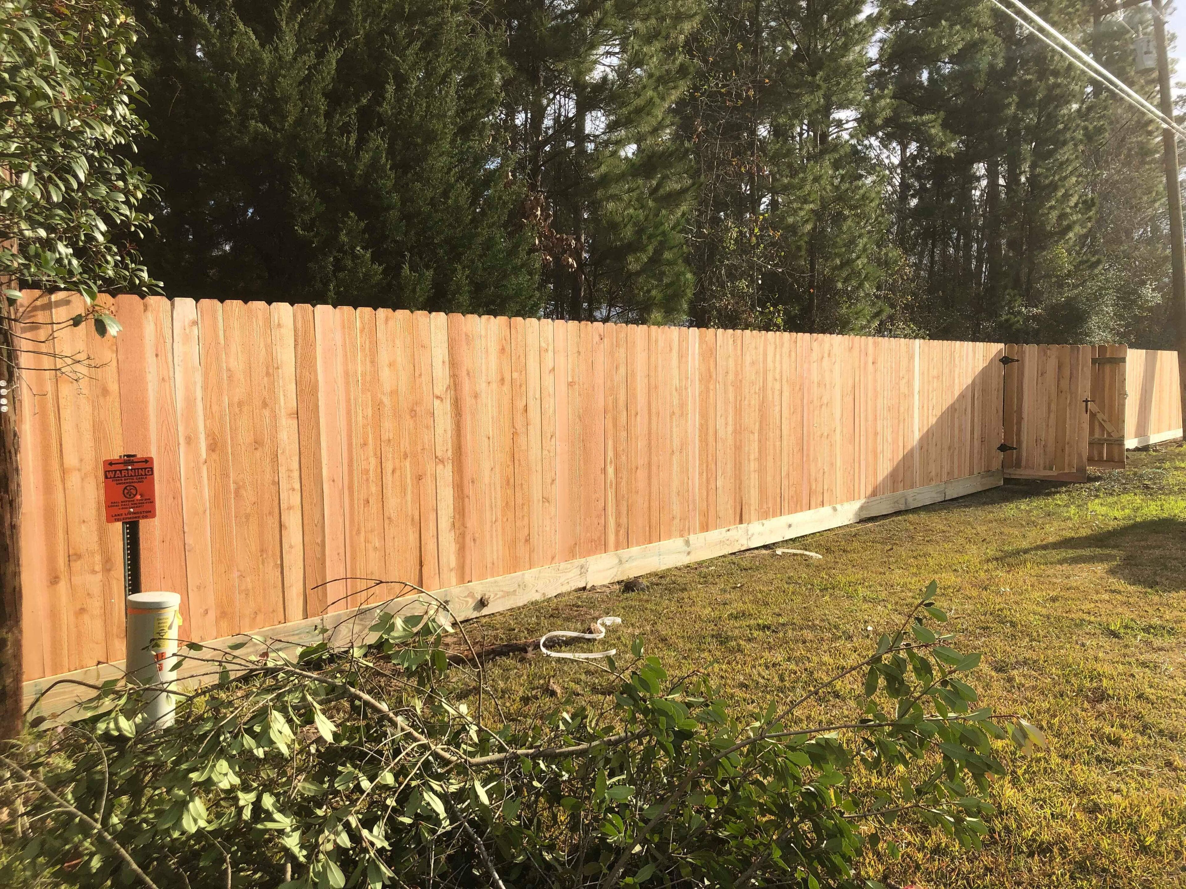 Een houten hek met een waarschuwingsbord, een betonnen paal en wat groenvoorziening ervoor.