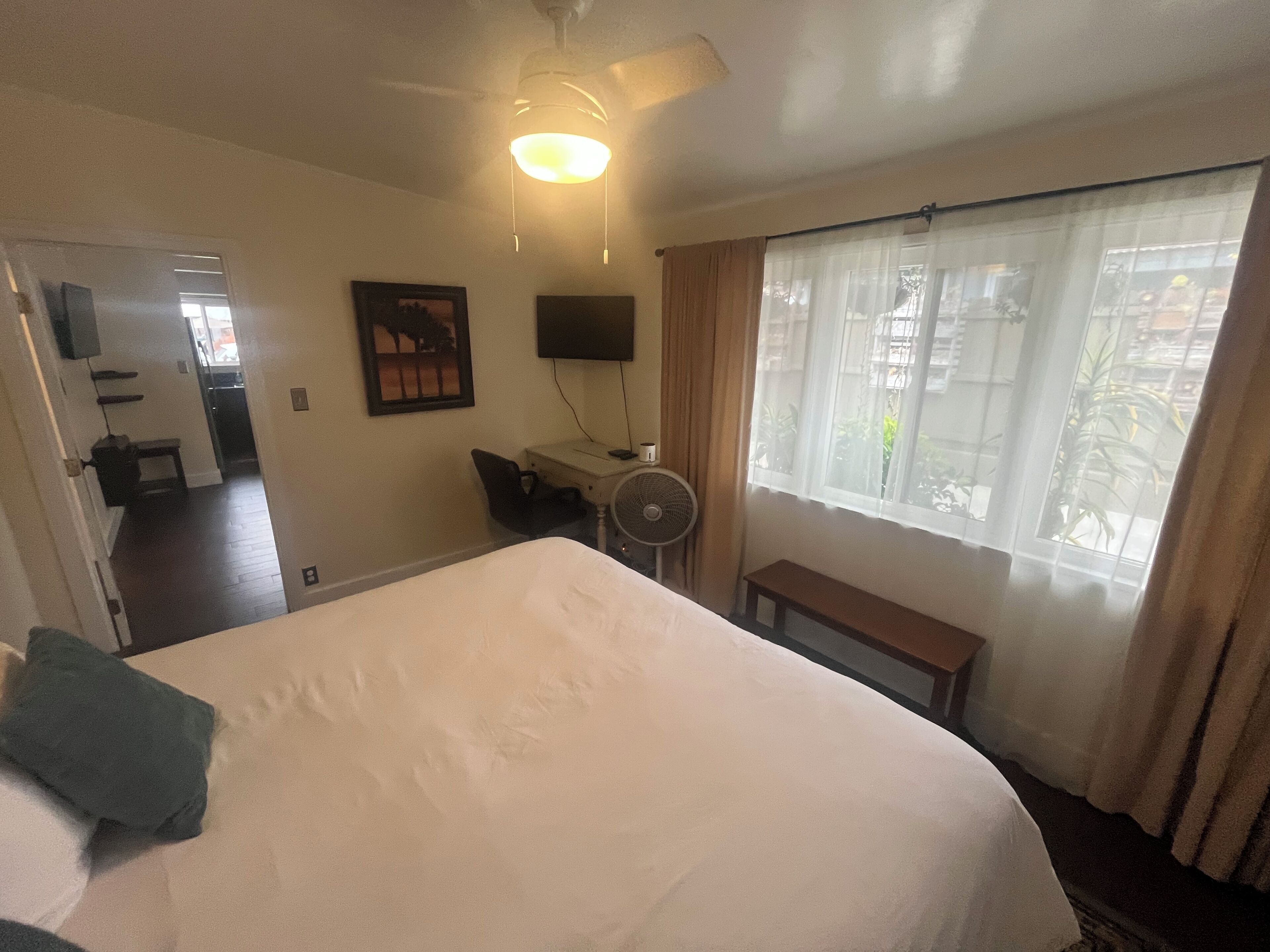 Una camera d'albergo con un letto grande, una scrivania con una sedia, una ventola, una TV e una finestra con tende.