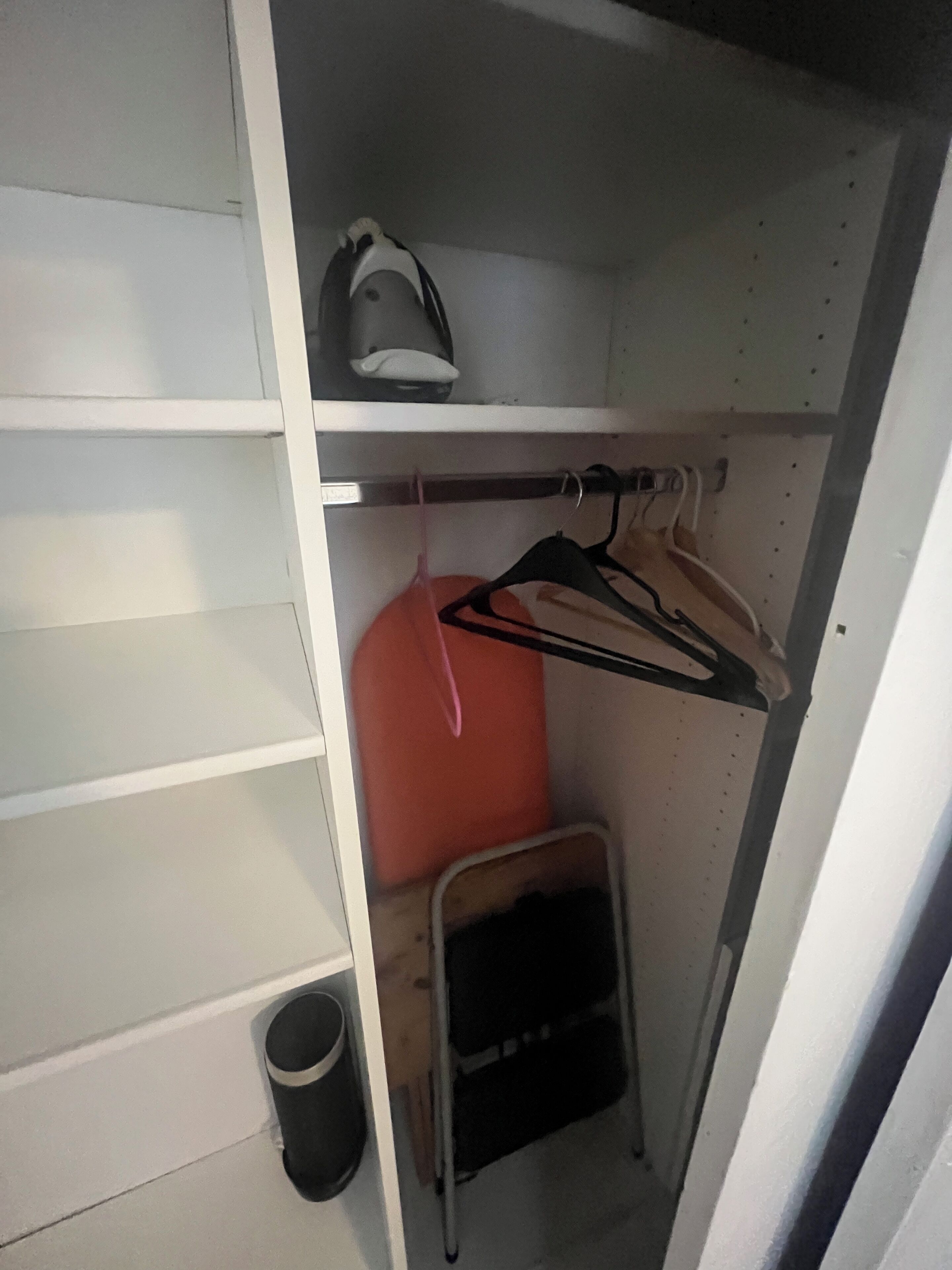 A small storage closet with shelves, a hanging rack, and a compact trash bin.