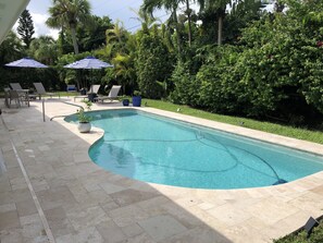 Una piscina rectangular con borde curvo, rodeada por una terraza con azulejos. Hay sillones de descanso y sombrillas, y el espacio está delimitado por una vegetación densa.